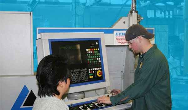 A manufacturing worker wearing a green shirt and a backwards black baseball hat demonstrating how to use a computer-aided machine to a woman with short dark hair wearing a white sweater.
