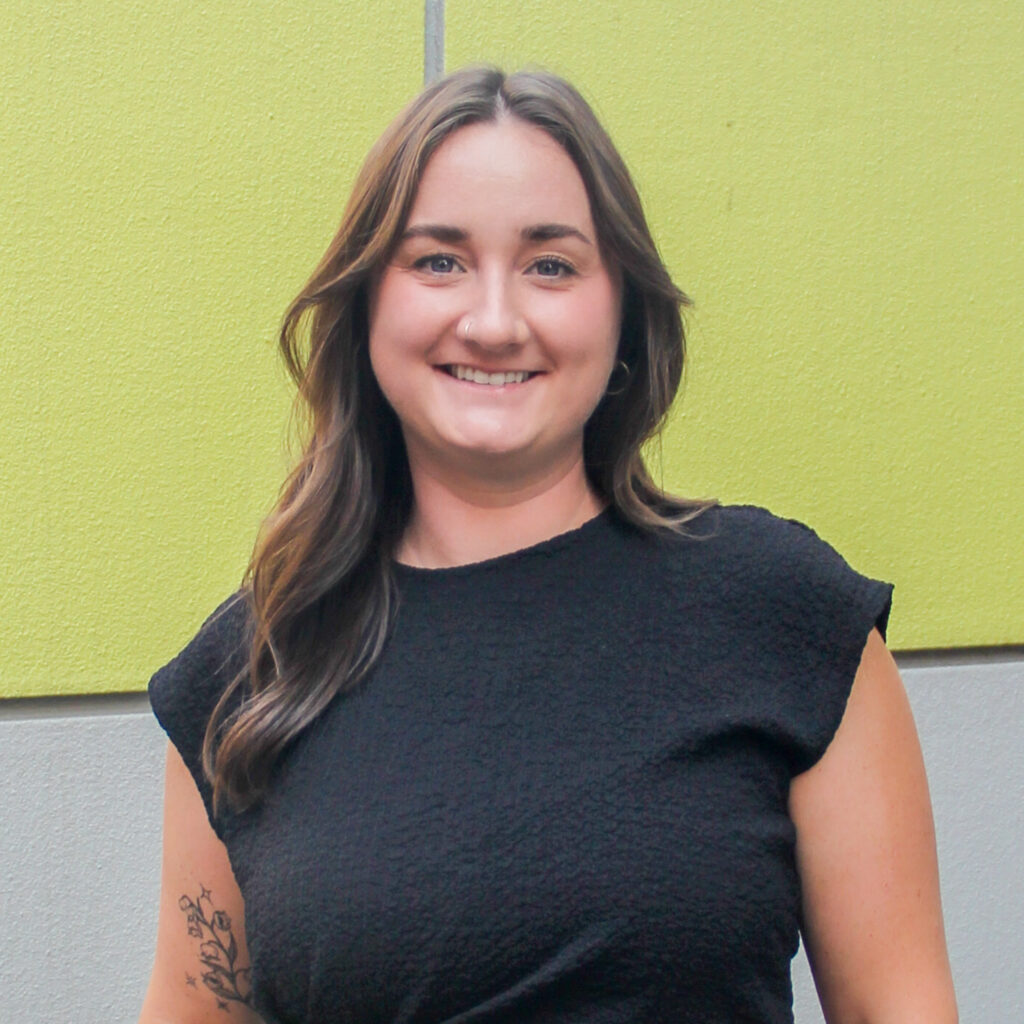 Portrait of a Caucasian woman with long brown hair wearing a black short-sleeve blouse standing in front of a green background.