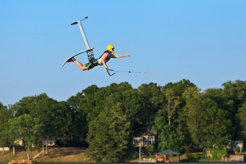 A man wearing a yellow helmet and black life jacket flying through the air after jumping on an Air Chair Hydrofoil watersport device.