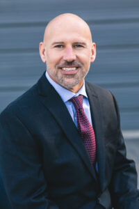 Portrait of a bald man with a short beard wearing a dark suit with a light blue collared shirt and burgundy and blue patterned tie.