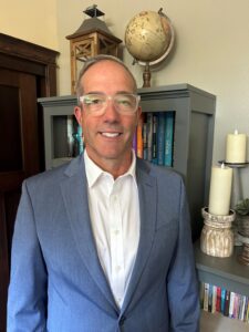 A portrait of a Caucasian man with short gray hair wearing transparent frame glasses, a slate blue sport jacket, and white collared shirt.