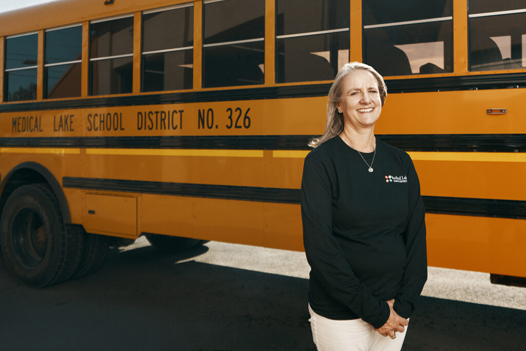 A Caucasian woman with dirty blonde shoulder-length hair wearing a black long-sleeve shirt and tan pants standing in front of a yellow school bus.