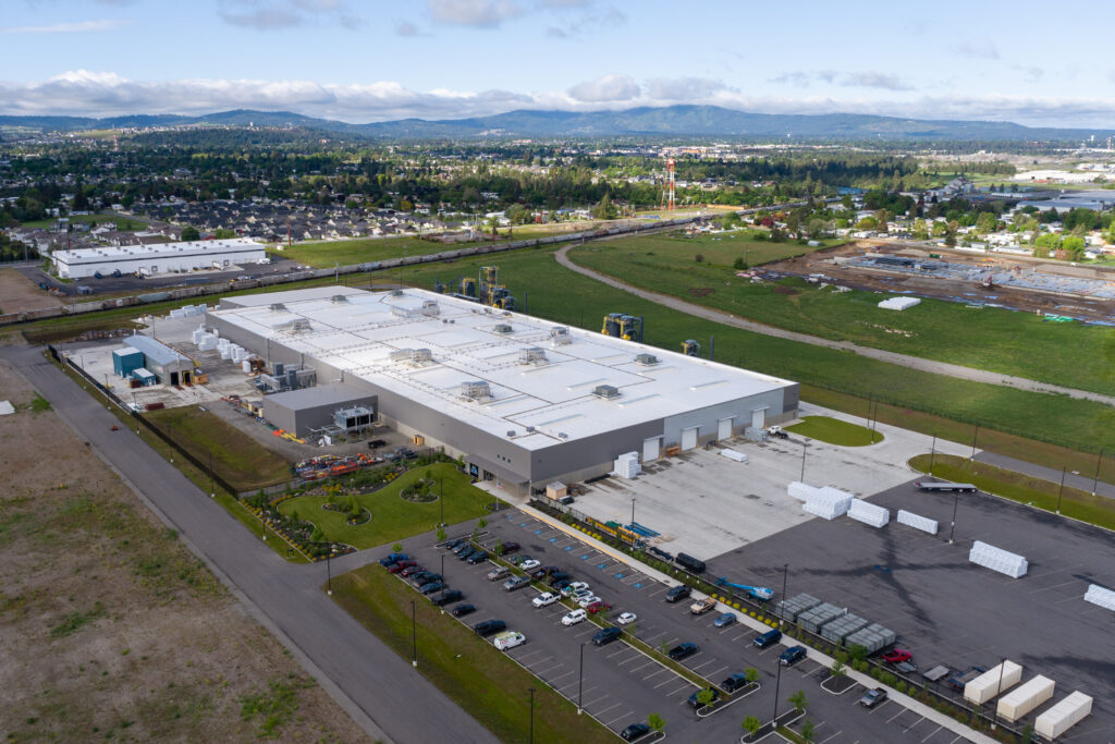 Aerial view of a large manufacturing facility with white-roofed buildings surrounded by a parking lot and green open space around it.