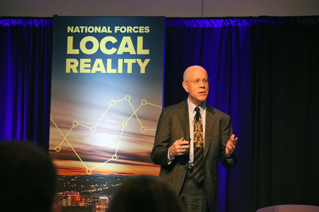 A bald Caucasian man with glasses wearing a gray suit and white dress shirt presenting on a stage in front of a display that says National Forces, Local Reality.