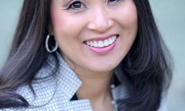 Alexa Allman Portrait of a woman with dark should-length hair wearing a white suit jacket with a black crisscross pattern on it over a black blouse.