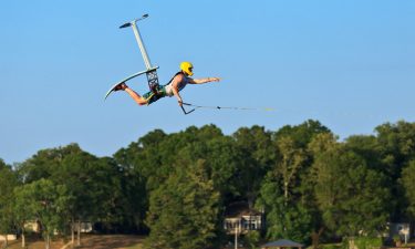 A man wearing a yellow helmet and black life jacket flying through the air after jumping on an Air Chair Hydrofoil watersport device.