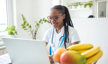 Portrait of young smiling female nutritionist in the consultation room. Nutritionist desk with healthy fruit, juice and measuring tape. Dietitian working on diet plan.