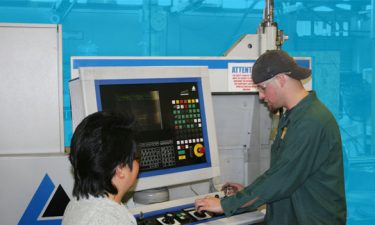 A manufacturing worker wearing a green shirt and a backwards black baseball hat demonstrating how to use a computer-aided machine to a woman with short dark hair wearing a white sweater.
